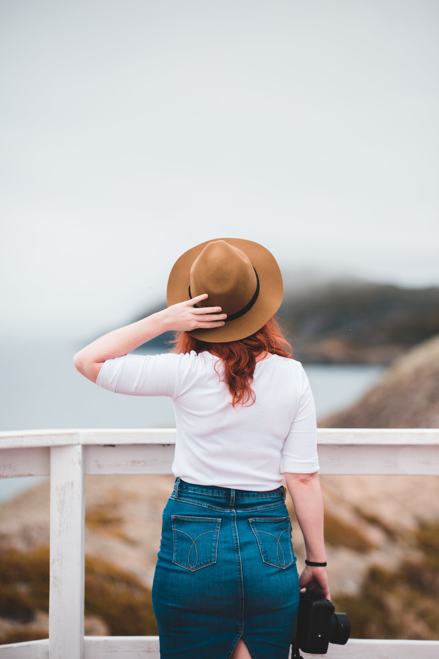 Back view faceless female traveler in casual outfit standing near waterfront railing and touching hat while enjoying views of blurred rough seashore