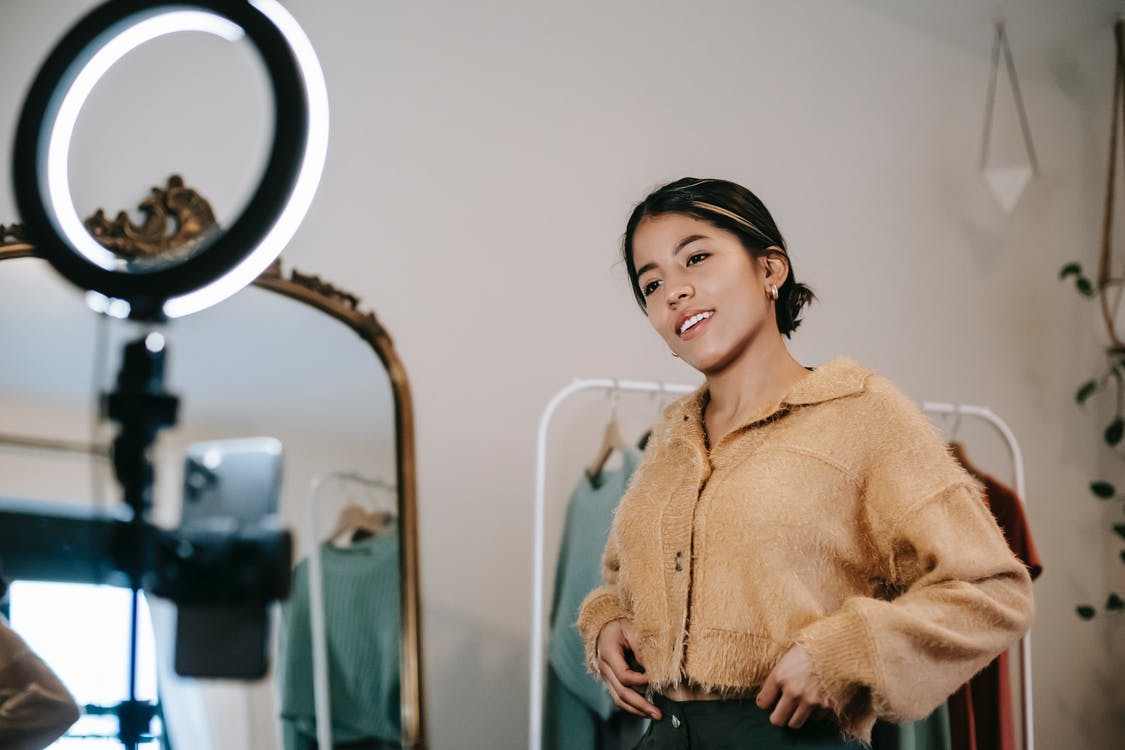 Low angle of positive ethnic female blogger shooting video on cellphone on tripod with shining lamp